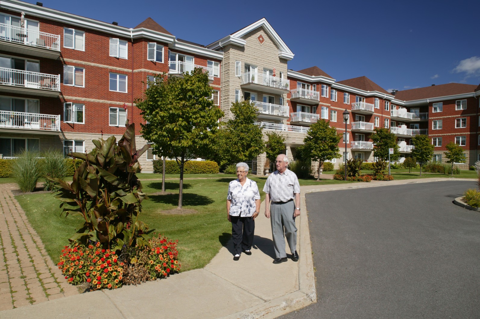 An elderly couple strolls together in front of a large red brick building, enjoying their time outdoors.