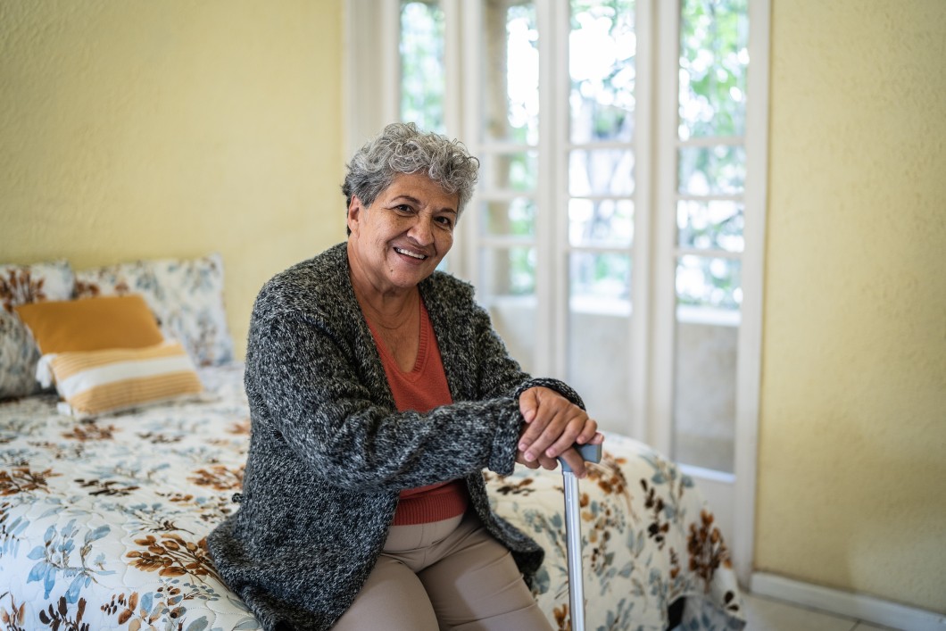 A woman sitting on a bed, holding a cane, with a smile on her face.