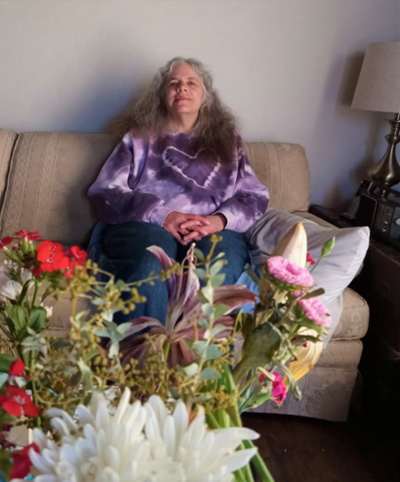 Older adult woman in purple tie-dye sweatshirt sitting on a couch with flowers on a table in front of her